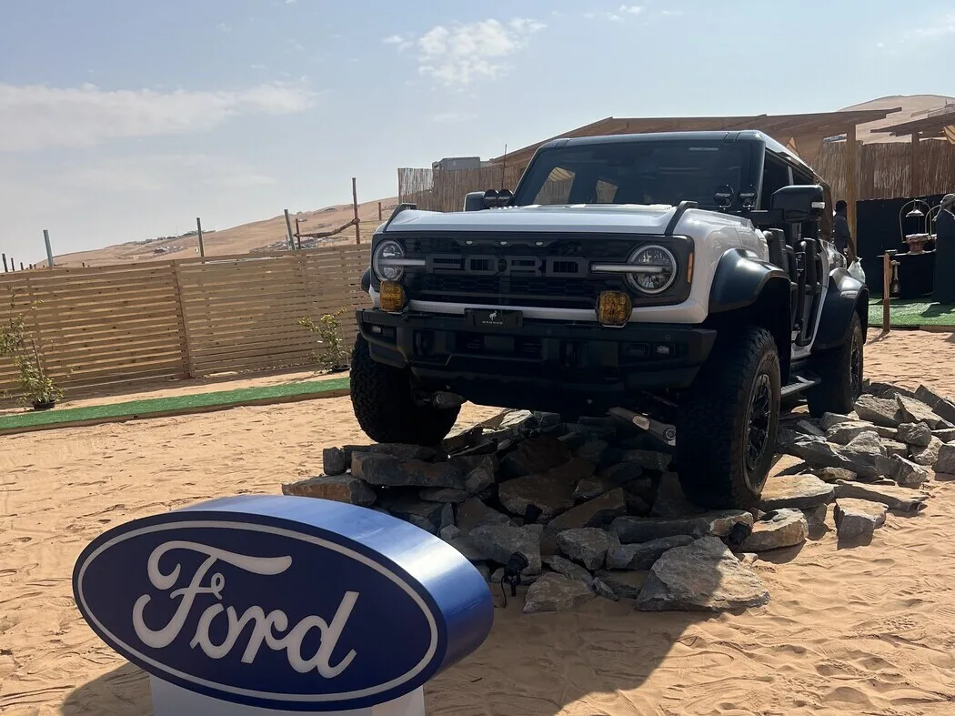 White Ford Bronco Raptor balanced on boulders beside a large Ford oval sign in the Liwa desert